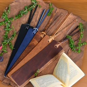 May include: A collection of leather bookmarks in various shades of brown, blue, and black, each with a leather cord. The bookmarks are arranged on a wooden surface with greenery and an open book.