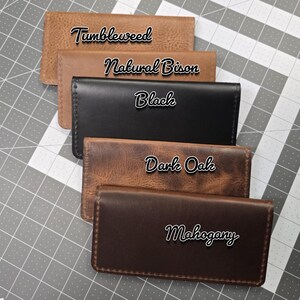 May include: Five leather wallets in different colors, stacked on top of each other. The colors are: Tumbleweed, Natural Bison, Black, Dark Oak, and Mahogany.