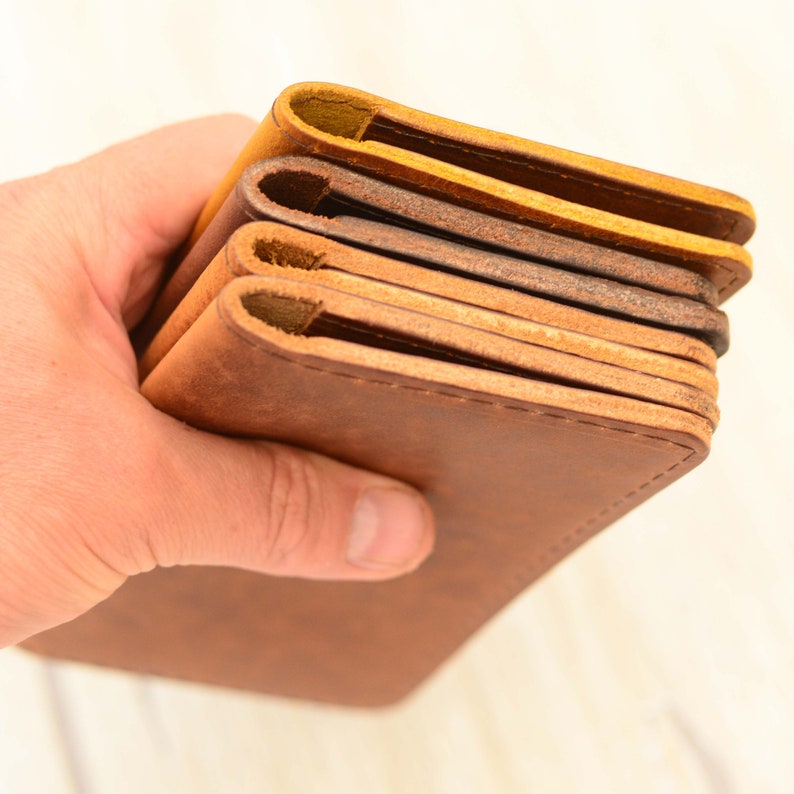 May include: A stack of four leather wallets in different shades of brown, from light to dark. The wallets are folded in half and have a stitched edge.