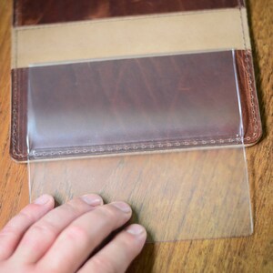 May include: A brown leather wallet with a clear plastic insert for displaying a business card or ID. The insert is partially pulled out of the wallet.