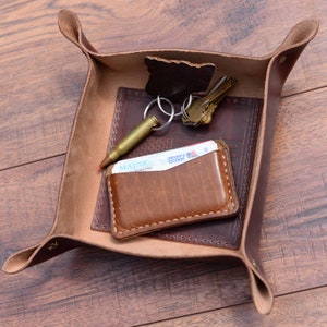 May include: A brown leather valet tray with a key ring, keys, a wallet, and a driver's license. The tray has a stitched edge and a decorative design.