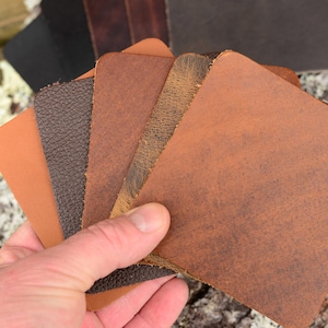 May include: A hand holds a fan of leather swatches in various shades of brown. The swatches display different textures, including smooth, pebbled, and distressed finishes. The leather colors range from light tan to dark brown.