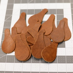 May include: A collection of brown leather teardrop-shaped cutouts. The cutouts are scattered across a gray and white grid surface. The leather has a natural, textured appearance, and the shapes vary slightly in size.