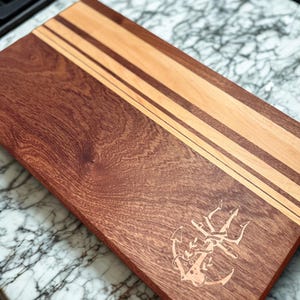 May include: A rectangular wooden cutting board with a dark reddish-brown base and lighter wood stripes. An etched anchor and trident design is visible on the board. The board is on a marble surface.