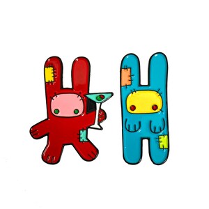 May include: Two cartoon bunny pins, one red with a martini glass and the other blue with a yellow face. Both have a patchwork design.