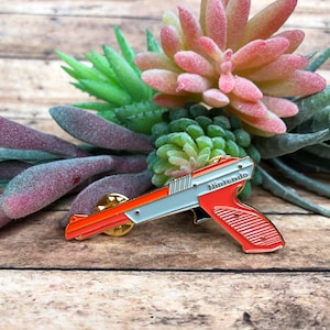 May include: Orange and white enamel pin of a Nintendo Zapper gun. The pin has a gold finish and the word "Nintendo" is printed on the side.