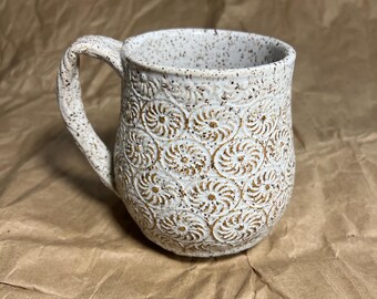 Puede incluir: Una taza de cerámica blanca moteada con un asa texturizada y un patrón de espiral repetido. La taza tiene una boca ancha y un cuerpo redondeado. El diseño es de color marrón cálido, contrastando con el fondo blanco.