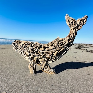 May include: A driftwood whale sculpture on a sandy beach under a clear blue sky. The whale is constructed from numerous pieces of weathered wood, showcasing a natural, rustic aesthetic. The sculpture's tail is raised, and it casts a shadow on the sand.
