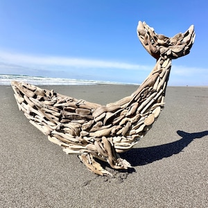 May include: A driftwood whale sculpture on a sandy beach under a blue sky. The whale is constructed from numerous pieces of weathered wood, with a curved body and a raised tail. The sculpture casts a shadow on the sand.