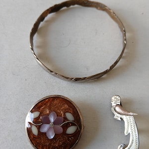 May include: A collection of jewelry consisting of a thin, textured metal bangle, a round brooch featuring a brown enamel background with a central light purple flower and white leaf inlays, and a silver-toned bird-shaped pin with engraved details.
