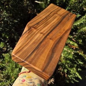 Può includere: Un tagliere rettangolare in legno con un motivo a venature naturali. La tavola è di colore marrone caldo e ha un bordo smussato. Il tagliere è mostrato ad angolo, con uno sfondo di foglie verdi.