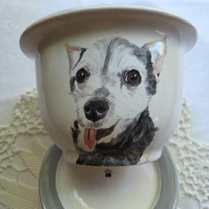 May include: A cream-colored ceramic planter featuring a hand-painted portrait of a black and white dog with brown eyes and a pink tongue sticking out. The planter sits on a matching cream-colored saucer with a gray rim and a small black paw print.