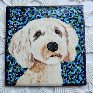 May include: A hand-painted ceramic tile featuring a portrait of a cream-colored dog with brown markings. The dog has a sweet expression and is surrounded by a blue, green, and black polka dot pattern.