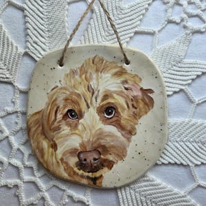 May include: A hand-painted ceramic wall plaque featuring a portrait of a golden-brown dog with shaggy fur and dark eyes. The off-white, speckled ceramic piece has rounded corners and is suspended by a thin, braided tan cord for hanging.