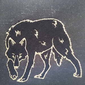 May include: A black silhouette of a wolf walking on a dark gray background. The wolf is outlined in gold thread.