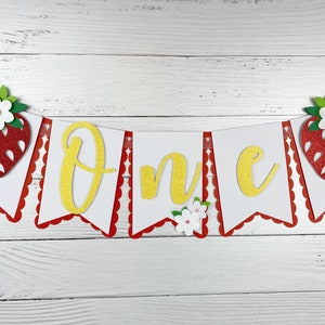 May include: A white and red banner with the word "One" in gold glitter letters. The banner is decorated with red and pink felt strawberries and white felt flowers.