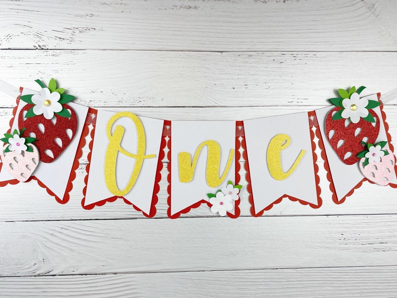 May include: A white and red banner with the word "One" in gold glitter letters. The banner is decorated with red and pink felt strawberries and white felt flowers.