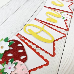 May include: A white banner with red scalloped edges and yellow glitter letters spelling "ONE". The banner is decorated with red and pink glitter strawberries and white flowers.