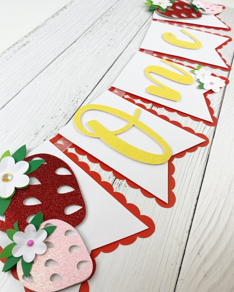 May include: A white banner with red scalloped edges and yellow glitter letters spelling "ONE". The banner is decorated with red and pink glitter strawberries and white flowers.