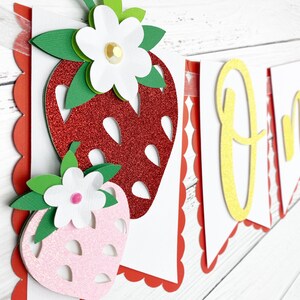 May include: A red and pink glitter strawberry banner with the word "One" in yellow glitter. The banner is made of paper and has a scalloped edge.