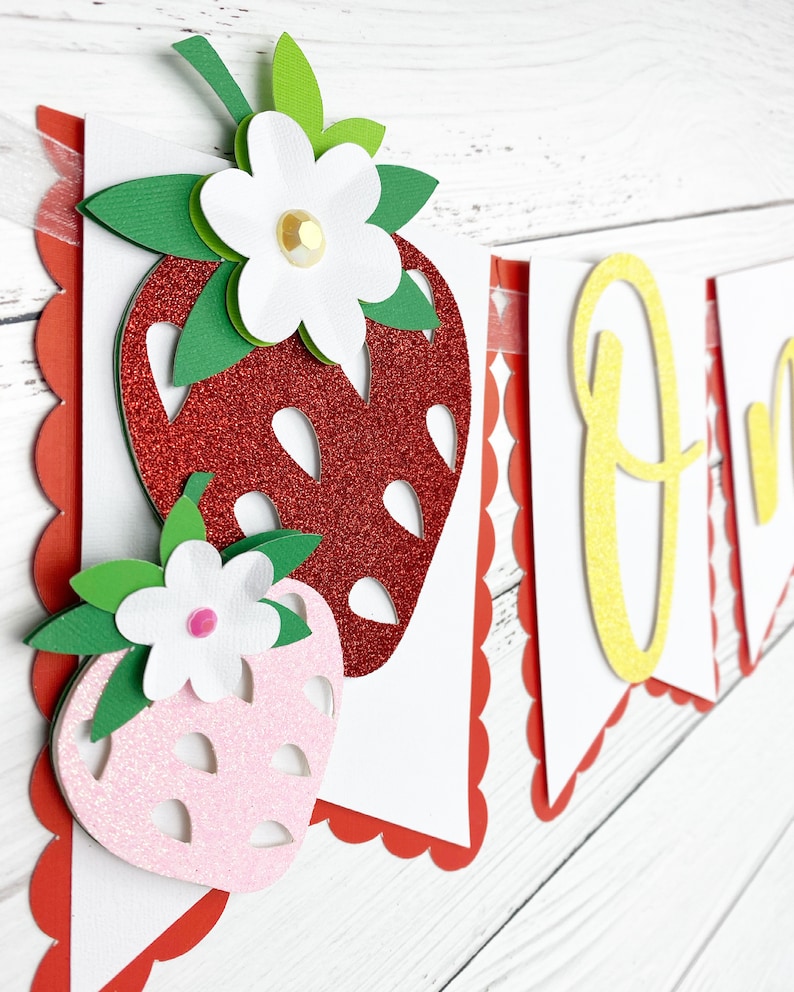 May include: A red and pink glitter strawberry banner with the word "One" in yellow glitter. The banner is made of paper and has a scalloped edge.