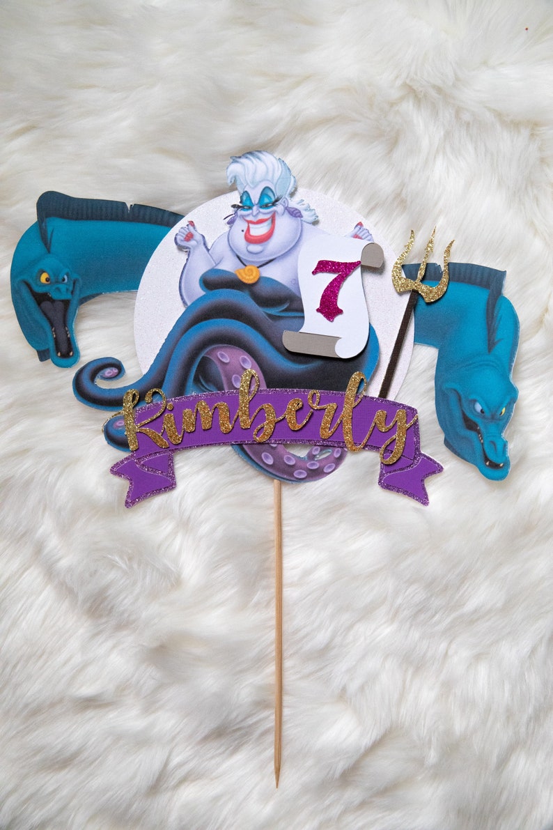May include: Purple and gold cake topper featuring Ursula from The Little Mermaid, a purple banner with the name Kimberly, and a scroll with the number 7.