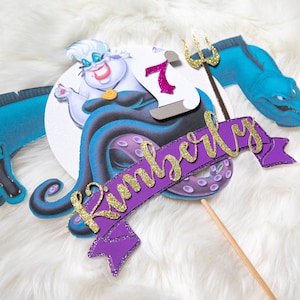 May include: Purple and gold cake topper featuring Ursula from The Little Mermaid, a purple banner with the name Kimberly, and a blue sea serpent.