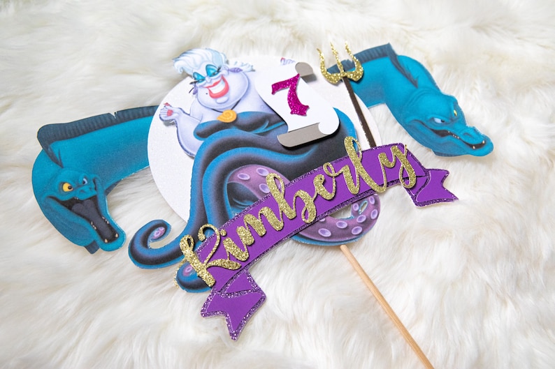 May include: Purple and gold cake topper featuring Ursula from The Little Mermaid, a purple banner with the name Kimberly, and a blue sea serpent.
