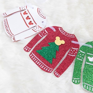 May include: A Christmas garland featuring three paper cutouts of sweaters with glitter. The sweaters are red, white, and green. The red sweater has a green Christmas tree and a yellow Mickey Mouse head. The white sweater has red hearts. The green sweater has a white Mickey Mouse head.