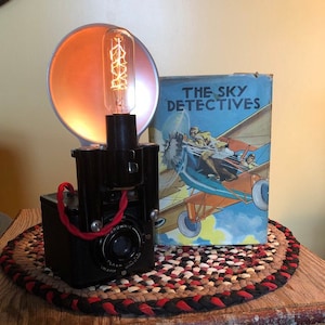 May include: A vintage black Brownie camera with a red strap and a flash attachment. The camera is sitting on a braided rug with a book titled "The Sky Detectives" behind it.