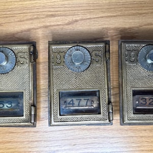May include: Three antique brass-toned post office box doors with combination dials and number displays. The boxes are rectangular with ornate detailing and display numbers 53, 477, and 326. The boxes are on a wooden surface.