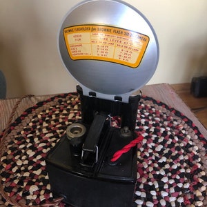 May include: A black Brownie Flash Six-20 camera with a silver flash attachment. The flash attachment has a chart with instructions for using the flash with different types of film and distances.