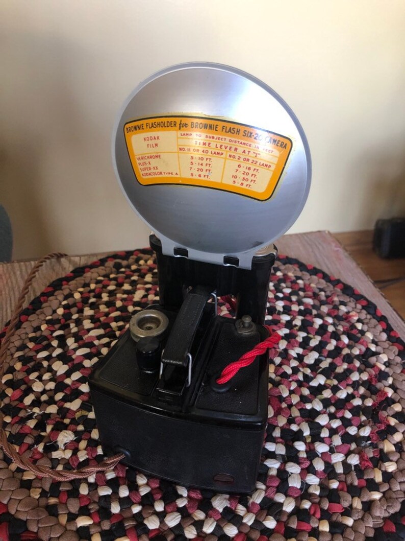 May include: A black Brownie Flash Six-20 camera with a silver flash attachment. The flash attachment has a chart with instructions for using the flash with different types of film and distances.