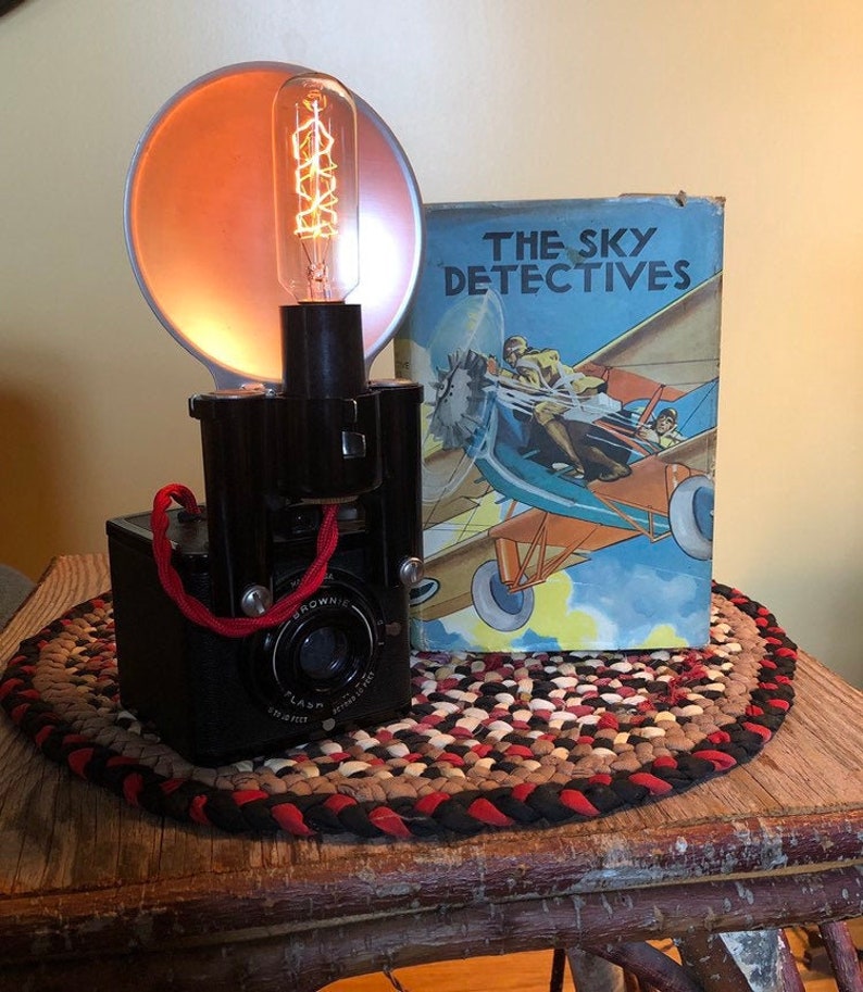 May include: A vintage black Brownie camera with a red strap and a flash attachment. The camera is sitting on a braided rug with a book titled "The Sky Detectives" behind it.