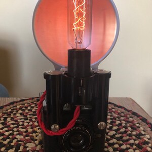 May include: A vintage black camera with a red cord and a light bulb attached to the top. The camera has a lens with the words "BROWNIE" and "6 TO 20 FEET" printed on it. The light bulb is a clear glass bulb with a coiled filament.