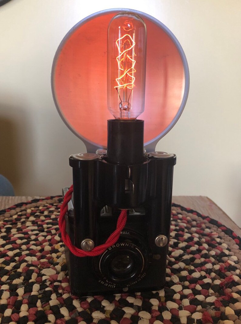 May include: A vintage black camera with a red cord and a light bulb attached to the top. The camera has a lens with the words "BROWNIE" and "6 TO 20 FEET" printed on it. The light bulb is a clear glass bulb with a coiled filament.
