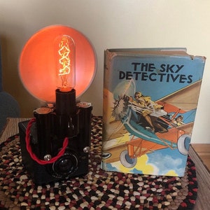 May include: A vintage camera lamp with a red cord and an orange lampshade. The lamp is sitting on a braided rug next to a book titled "The Sky Detectives".