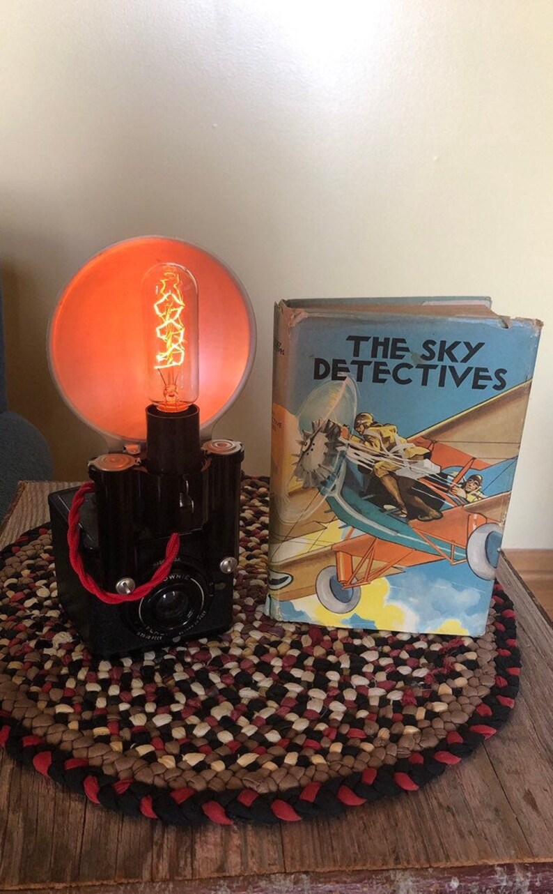 May include: A vintage camera lamp with a red cord and an orange lampshade. The lamp is sitting on a braided rug next to a book titled "The Sky Detectives".
