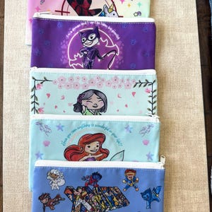 Może przedstawiać: Stos pięciu pi&oacute;rnik&oacute;w zapinanych na zamek błyskawiczny z kolorowymi wzorami. Pi&oacute;rniki przedstawiają ilustracje postaci z komiks&oacute;w i animacji, w tym Deadpoola, Catwoman, Ariel i postaci X-Men. Pi&oacute;rniki są w r&oacute;żnych kolorach, w tym r&oacute;żowym, fioletowym i niebieskim.