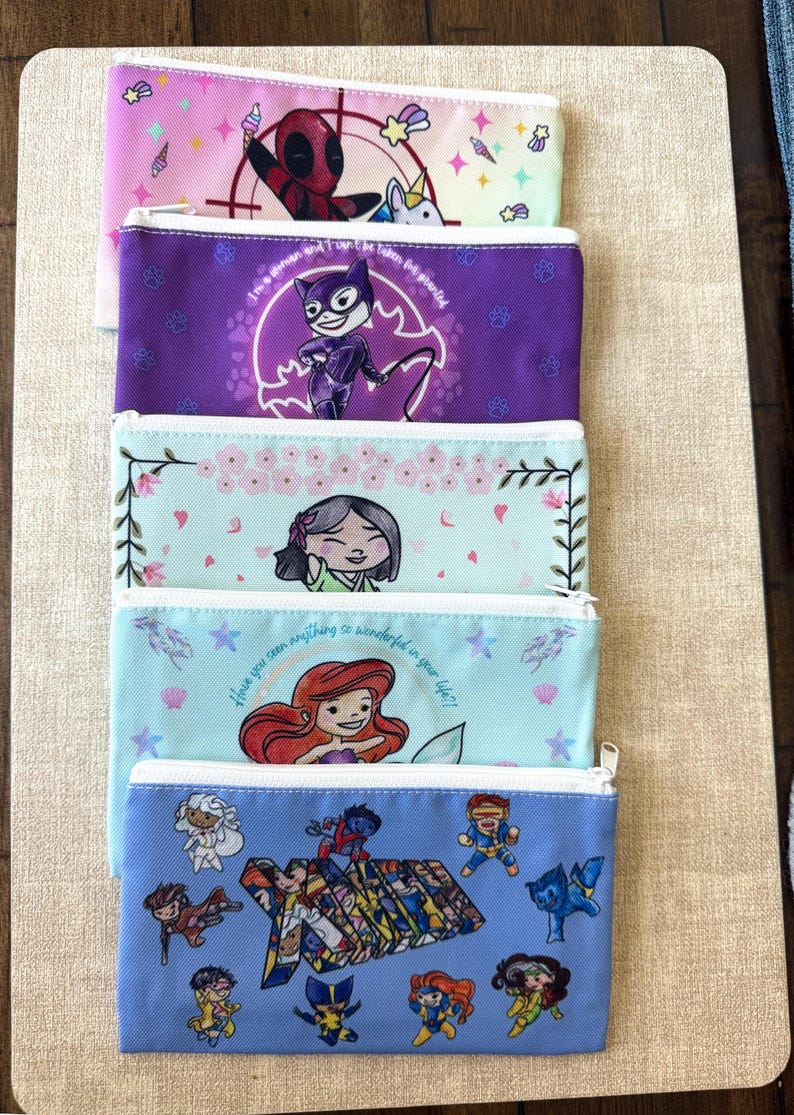 Może przedstawiać: Stos pięciu pi&oacute;rnik&oacute;w zapinanych na zamek błyskawiczny z kolorowymi wzorami. Pi&oacute;rniki przedstawiają ilustracje postaci z komiks&oacute;w i animacji, w tym Deadpoola, Catwoman, Ariel i postaci X-Men. Pi&oacute;rniki są w r&oacute;żnych kolorach, w tym r&oacute;żowym, fioletowym i niebieskim.