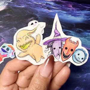 Puede incluir: Una colección de pegatinas coloridas con personajes de "Pesadilla antes de Navidad". Las pegatinas incluyen a Oogie Boogie, una bruja, un personaje diabólico y una calavera. Las pegatinas se sostienen en una mano sobre un fondo oscuro y texturizado.