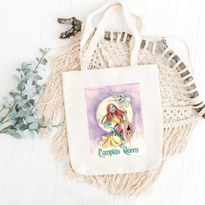 May include: A cream-coloured tote bag with a watercolour illustration of a character with red hair, a patchwork dress, and a ghost. The text "Pumpkin Queen" is below the image. The bag has cream-coloured handles.