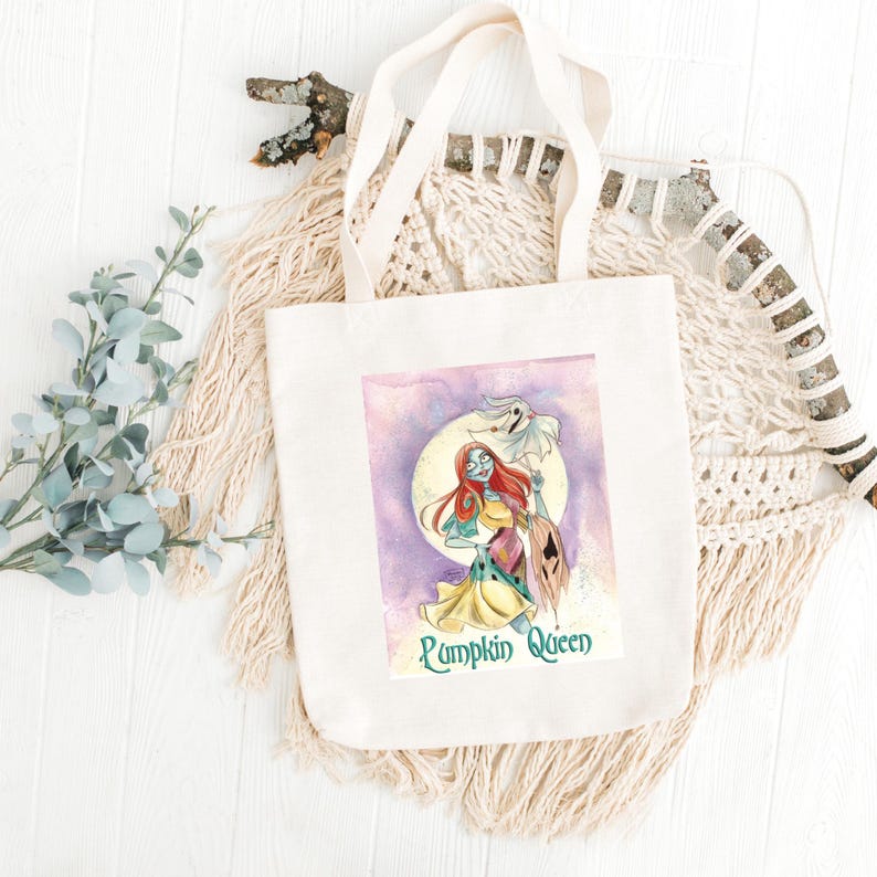 May include: A cream-coloured tote bag with a watercolour illustration of a character with red hair, a patchwork dress, and a ghost. The text "Pumpkin Queen" is below the image. The bag has cream-coloured handles.