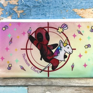 Może przedstawiać: Prostokątny pi&oacute;rnik z zapięciem na zamek błyskawiczny. Projekt przedstawia postać z kresk&oacute;wki Deadpoola jadącego na jednorożcu, otoczonego gwiazdami, rożkami lod&oacute;w i tekstem "Maximum Effort!" na gradientowym tle.