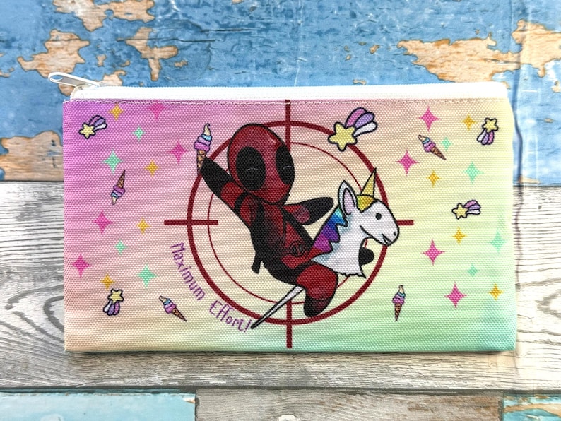 Może przedstawiać: Prostokątny pi&oacute;rnik z zapięciem na zamek błyskawiczny. Projekt przedstawia postać z kresk&oacute;wki Deadpoola jadącego na jednorożcu, otoczonego gwiazdami, rożkami lod&oacute;w i tekstem "Maximum Effort!" na gradientowym tle.