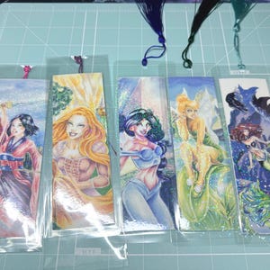 May include: Five rectangular bookmarks in clear plastic sleeves, each featuring a colorful illustration of a different character. The bookmarks include tassels in blue, black, and green, and display artwork of various figures with glittery accents.