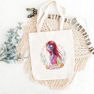 May include: A cream-coloured canvas tote bag with a colourful illustration of a character with red hair and a floral wreath. The character is surrounded by a floral wreath with the word "Queen" at the bottom. The bag has cream-coloured handles.