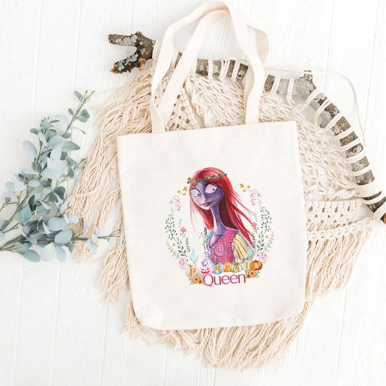 May include: A cream-coloured canvas tote bag with a colourful illustration of a character with red hair and a floral wreath. The character is surrounded by a floral wreath with the word "Queen" at the bottom. The bag has cream-coloured handles.