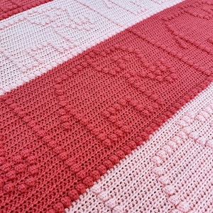 May include: Close-up of a handmade crochet blanket with alternating stripes of red and light pink. The red stripes feature a textured, raised stitch pattern, while the pink stripes have a simpler design. The blanket is made of soft yarn.