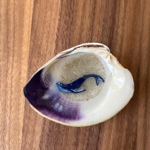 Op de afbeelding: Een decoratieve zeeschelp met een paars omrande binnenkant, gevuld met zand en een klein blauw walvisfiguur. De schelp staat tegen een houten achtergrond, wat een nautisch thema creëert.